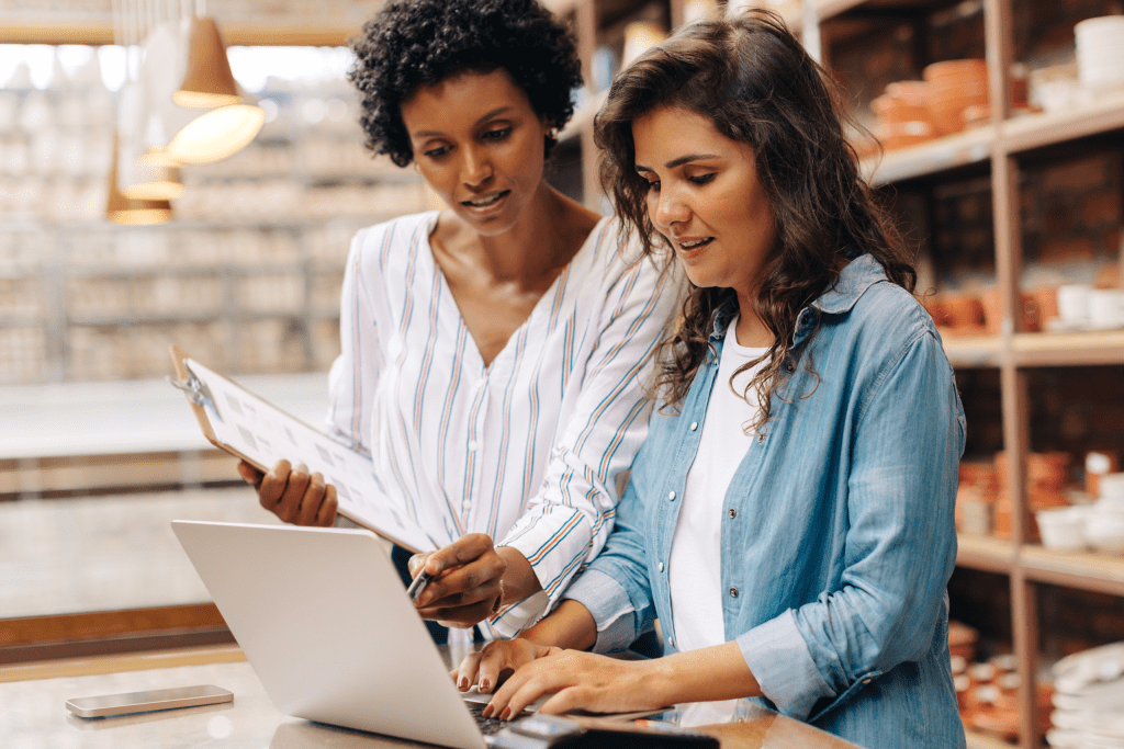 Diverse pair of women entrepreneurs working in their pottery store while using a laptop